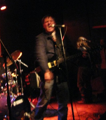 Man on dark stage in front of drum set, strumming guitar and yelling into microphone