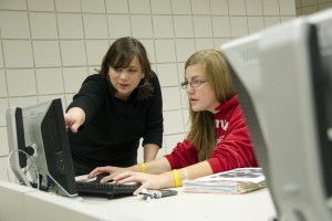 Student working at a computer station with instructor leaning over and pointing.